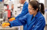 Woman in blue lab coat and safety goggles placing a sample on machine 